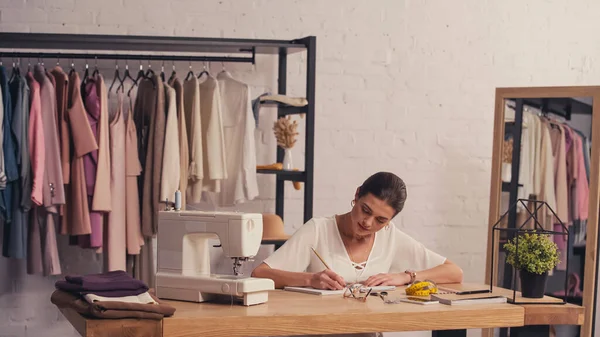 Diseñador escribiendo en cuaderno cerca de tela y máquina de coser en atelier - foto de stock