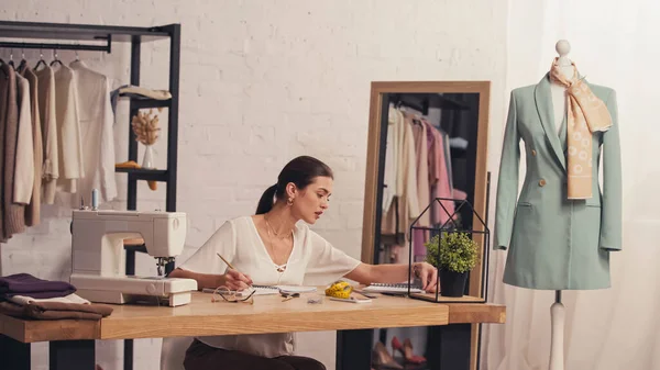 Brünette Designerin schaut im Atelier neben Nähmaschine und Stoff auf Notizbuch — Stockfoto