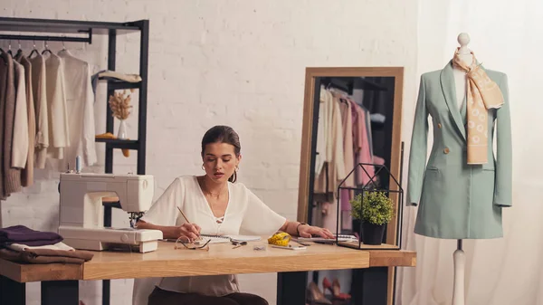 Brunette seamstress writing on notebook near mannequin and sewing machine in atelier — Stock Photo