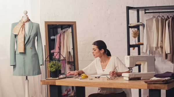 Näherin arbeitet mit Notizbüchern in der Nähe von Nähmaschine und Stoff im Atelier — Stockfoto