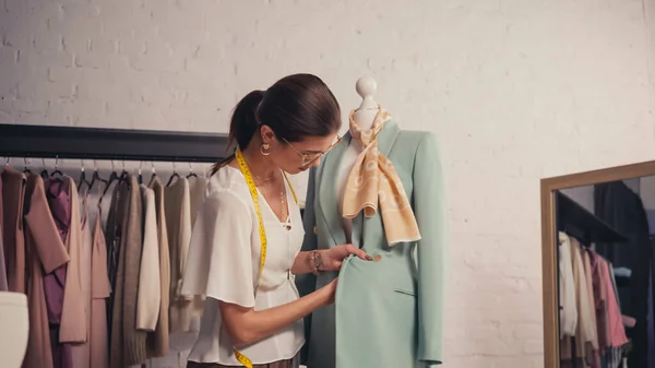 Brunette designer avec ruban à mesurer de travail avec blazer sur mannequin en atelier — Photo de stock