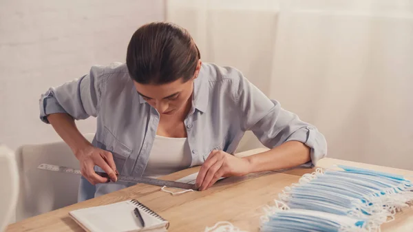 Brunette designer measuring medical mask near notebook in atelier — Stock Photo