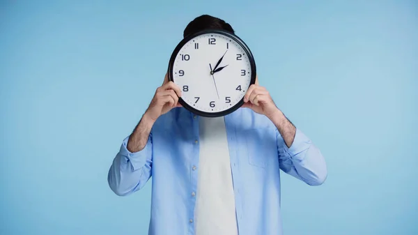 Man in shirt holding clock while covering face isolated on blue background — Stock Photo