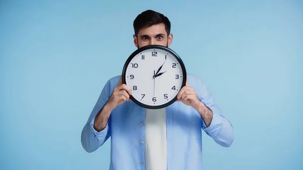 Man in shirt holding clock while covering face and looking at camera isolated on blue — Stock Photo