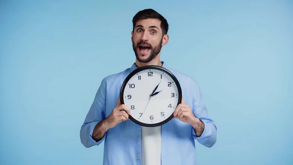 Homme barbu en chemise tenant horloge tout en regardant la caméra isolée sur fond bleu — Photo de stock