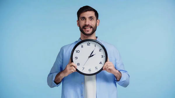 Uomo stordito in camicia che tiene orologio mentre guarda la fotocamera isolata su sfondo blu — Foto stock