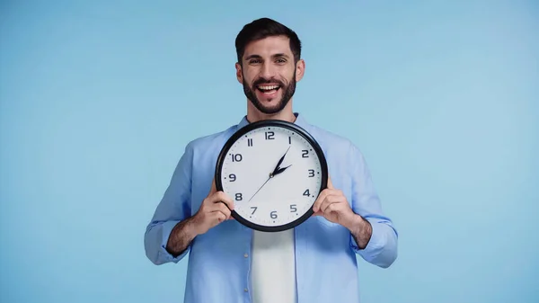 Fröhlicher Mann im Hemd, Uhr in der Hand, während er isoliert in die Kamera auf blauem Hintergrund blickt — Stockfoto