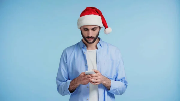 Barbudo hombre en rojo santa sombrero de mensajería en el teléfono inteligente aislado en azul - foto de stock
