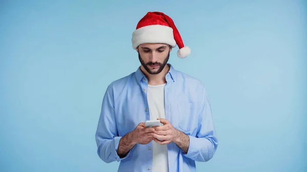 Barbudo homem no vermelho santa chapéu mensagens no smartphone isolado no azul — Fotografia de Stock