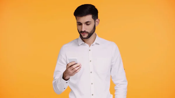 Hombre barbudo en camisa blanca mirando teléfono inteligente aislado en amarillo - foto de stock