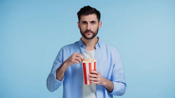 Бородатий чоловік тримає смугасте відро з попкорном ізольовано на синьому — стокове фото