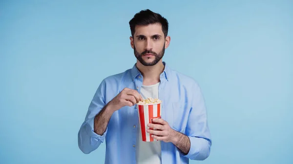 Barbudo hombre sosteniendo cubo rayado con palomitas de maíz aislado en azul - foto de stock