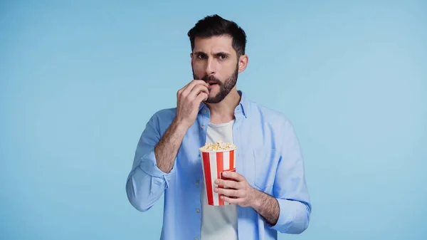 Bärtiger Mann hält roten Eimer und isst Popcorn isoliert auf blauem Grund — Stockfoto