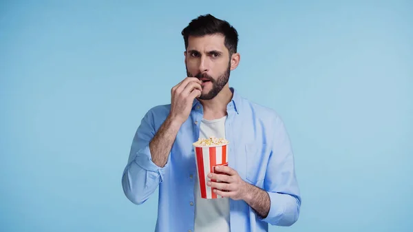 Homem barbudo segurando balde vermelho e comer pipocas isoladas em azul — Fotografia de Stock