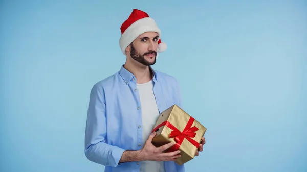Bärtiger Mann mit roter Weihnachtsmütze und Weihnachtsgeschenk isoliert auf blauem Grund — Stockfoto