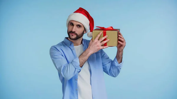 Curioso barbudo hombre en rojo santa sombrero sacudiendo navidad presente aislado en azul - foto de stock