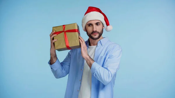 Curious man in red santa hat shaking christmas present isolated on blue — Stock Photo