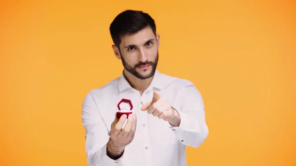 Homme barbu en chemise blanche tenant boîte avec bague de fiançailles tout en pointant du doigt la caméra isolée sur jaune — Photo de stock