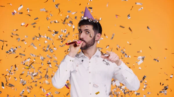Bearded man in white shirt blowing in party horn near falling confetti on yellow — Stock Photo