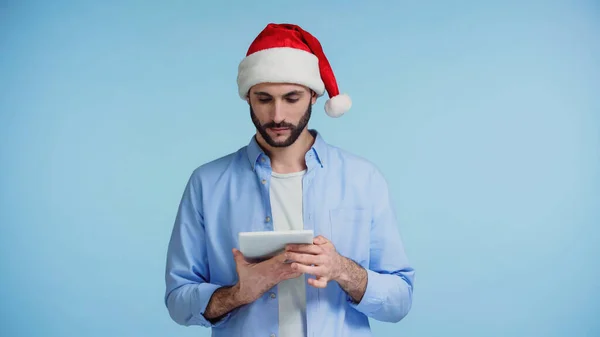 Hombre barbudo en sombrero de santa rojo usando tableta digital aislado en azul - foto de stock