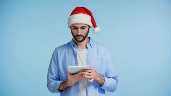 Homem barbudo em chapéu vermelho santa usando tablet digital isolado em azul — Fotografia de Stock