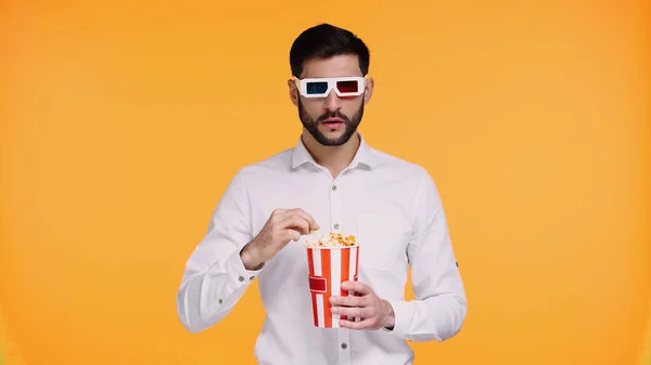 Bärtiger Mann in 3D-Brille hält gestreiften Eimer mit Popcorn in der Hand und schaut Film isoliert auf gelb — Stockfoto