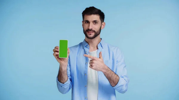 Hombre feliz en camisa apuntando con el dedo al teléfono inteligente con pantalla verde aislado en azul - foto de stock