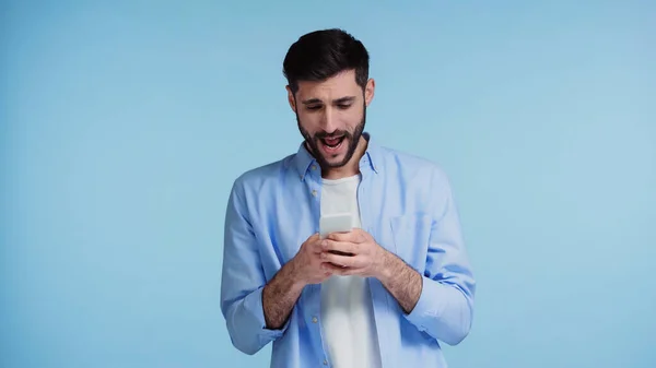 Homem barbudo usando smartphone e gritando enquanto mensagens isoladas em azul — Fotografia de Stock
