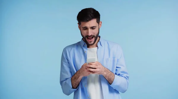 Hombre barbudo usando teléfono inteligente y gritando mientras mensajería aislado en azul — Stock Photo