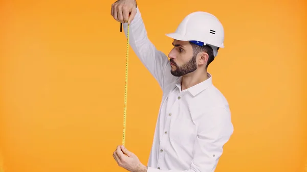 Hombre enfocado en casco de seguridad mirando cinta métrica aislada en amarillo - foto de stock