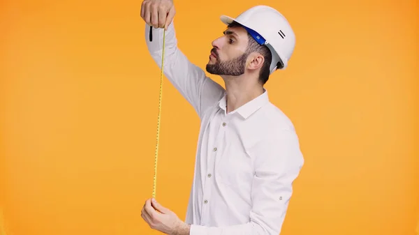 Bearded engineer in safety helmet looking at measuring tape isolated on yellow — Stock Photo