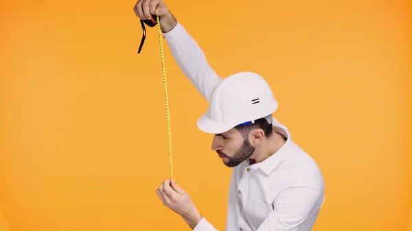 Architecte concentré dans le casque de sécurité regardant ruban de mesure isolé sur jaune — Photo de stock