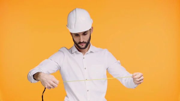 Konzentrierter Ingenieur mit harter Mütze, der auf gelb isoliertes Maßband blickt — Stockfoto