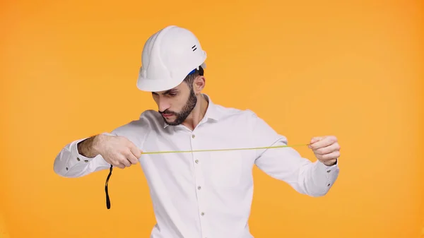 Ingeniero barbudo en sombrero duro mirando cinta métrica aislada en amarillo - foto de stock