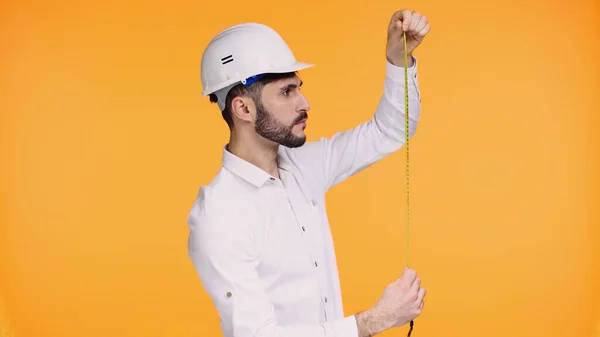 Bearded architect in hard hat looking at measuring tape isolated on yellow — Stock Photo