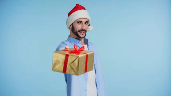 Bearded man in red santa hat giving wrapped christmas present isolated on blue — Stock Photo
