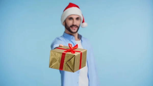 Hombre feliz en sombrero de santa rojo dando regalo de Navidad envuelto aislado en azul - foto de stock