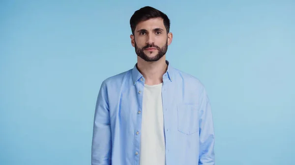 Hombre confiado y barbudo en camisa mirando a la cámara mientras está de pie aislado en azul - foto de stock