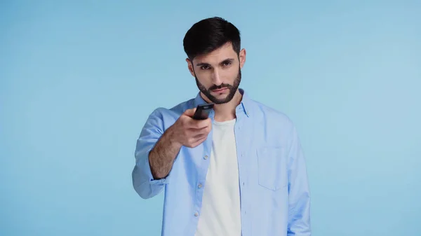 Hombre barbudo mirando a la cámara mientras sostiene el mando a distancia y haciendo clic en canales aislados en azul - foto de stock