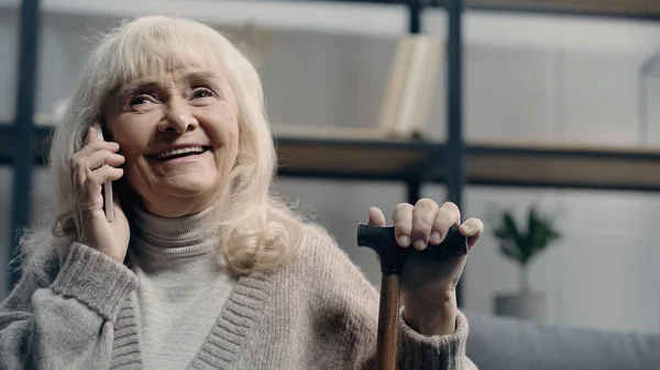 Fröhliche Seniorin spricht mit Smartphone und hält Gehstock in der Hand — Stockfoto