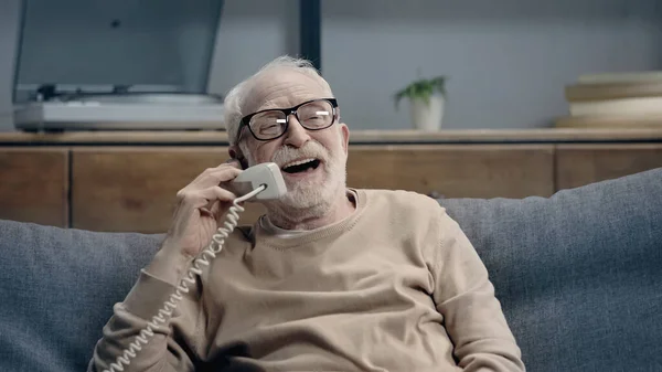 Homem idoso alegre em óculos falando no telefone vintage e sentado no sofá em casa — Fotografia de Stock