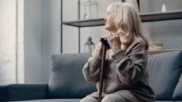 Senior woman with dementia sitting on sofa with walking stick — Stock Photo