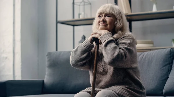 Sad senior woman with dementia sitting on sofa while leaning on walking cane — Stock Photo