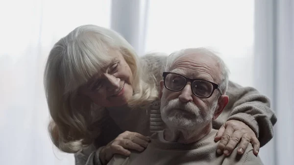 Cheerful senior woman calming down husband in eyeglasses with dementia — Stock Photo