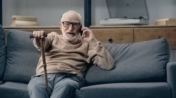 Heureux homme âgé parlant sur smartphone et assis sur le canapé avec canne à pied — Photo de stock