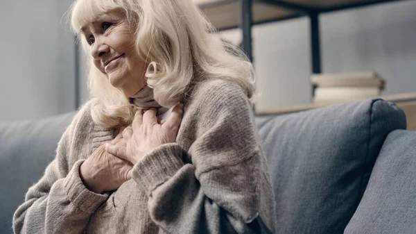 Pleased senior woman in sweater holding photograph near chest at home — Stock Photo