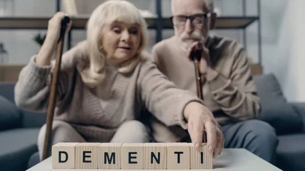 Senior man sitting near woman putting wooden cube with lettering and creating word dementia — Stock Photo