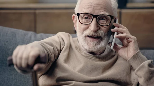 Hombre mayor barbudo en gafas graduadas hablando en el teléfono inteligente en casa - foto de stock