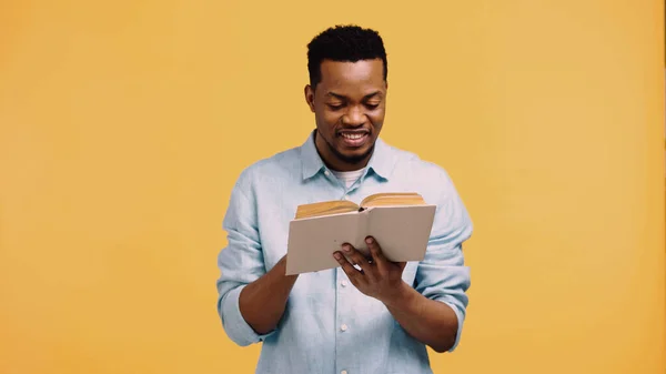Happy african american man in blue shirt reading book isolated on yellow — Stock Photo
