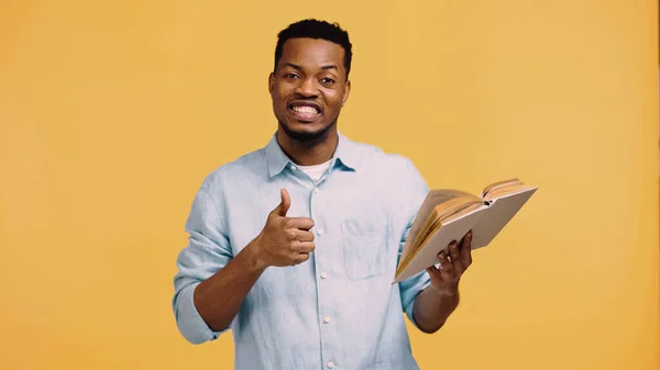 Happy african american man in blue shirt showing thumb up and holding book isolated on yellow — Stock Photo